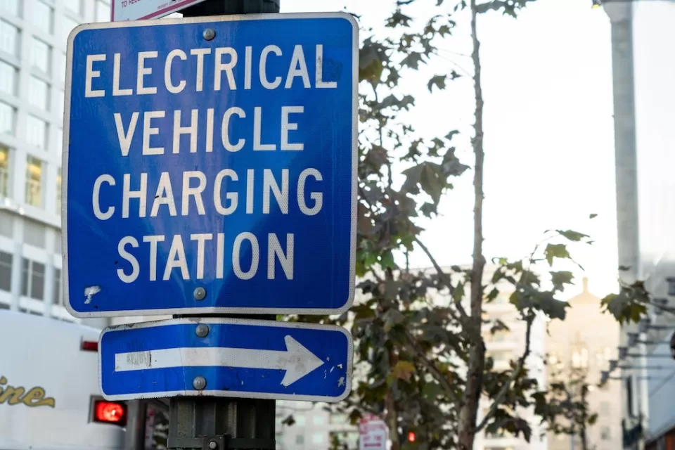 Close-up on blue and white "Electric vehicle charging station" sign with arrow pointing right on pole on city street.